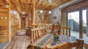 Un intérieur rustique en bois, avec un coin repas, des chaises en bois, une table recouverte d’une nappe à motifs et une grande fenêtre offrant une vue panoramique.