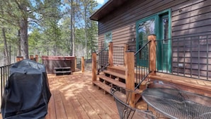 Una terraza de madera con una parrilla, un jacuzzi y una escalera que lleva a una cabaña con detalles verdes.