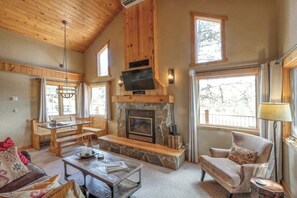 A cozy living room with a stone fireplace, wooden ceiling, and large windows. - Serenity Ridge Retreat – Cozy Cabin with Hot Tub & Mountain Views Near Terry Peak (Lead)