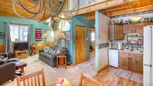 A spacious living room with a large chandelier, a cozy sofa, and a television. The kitchen features wooden cabinets and a microwave. - Cozy Black Hills Retreat – Loft, deck, pet-friendly & peaceful mountain views! (Lead)