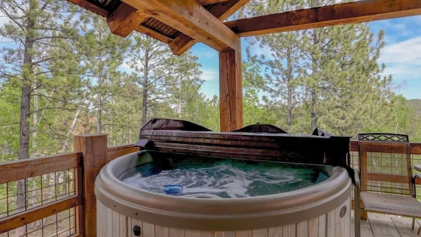 A wooden deck with a hot tub, surrounded by a wooden railing and trees. - Mercantile Cabin (Lead)