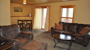A living room with a brown leather couch, a matching ottoman, and a small table. There is a window with curtains, a door, and a wall-mounted light fixture. - Mercantile Cabin (Lead)