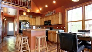 A kitchen with wooden cabinets, a central island with stools, a dining table with chairs, and large windows. - Mercantile Cabin (Lead)