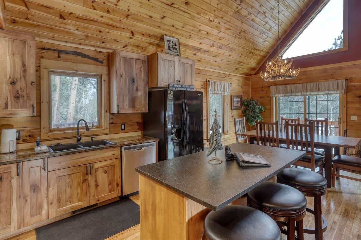 A wooden cabin interior with a kitchen, dining area, and a large window overlooking the forest.