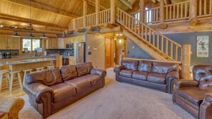 A spacious living area with a high ceiling and wooden beams, featuring a leather sofa, a leather armchair, and a staircase leading to an upper level. - Deer Haven Lodge – Spacious Family Cabin with Hot Tub, Game Room & Scenic Views (Lead)