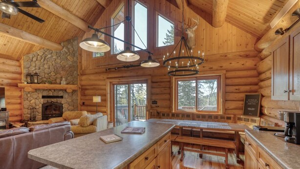 A log cabin interior with a stone fireplace, wooden dining area, and large windows. - Deer Haven Lodge – Spacious Family Cabin with Hot Tub, Game Room & Scenic Views (Lead)