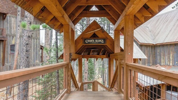 Eine Holzbrücke führt zu einem Schild des Cash's Slushie Lodge, umgeben von Bäumen und rustikalen Gebäuden.