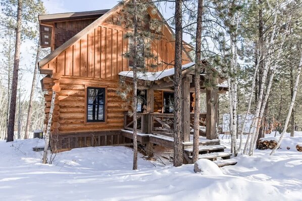 Une cabane en bois dotée d’un porche et d’un escalier, entourée d’arbres et de neige.