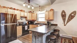 A modern kitchen with wooden cabinets, a stainless steel refrigerator, a central island with stools, and a hanging light fixture. - Lookout View Retreat – Cozy 3-Level Cabin with Hot Tub, Loft & Mountain Views (Lead)