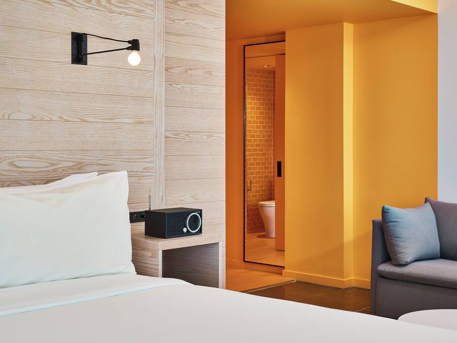A modern hotel room with a wooden wall, a bedside table with a radio, a sofa, and a bathroom visible through an open door.