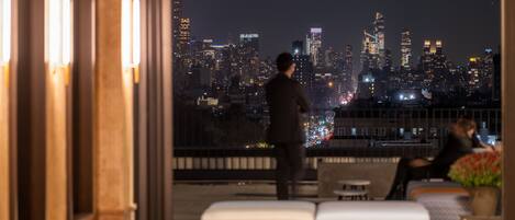 Ein modernes Hotelzimmer mit Stadtblick, einem Balkon inklusive Sitzgelegenheit und einem großen Fenster zum Blick auf den Horizont.