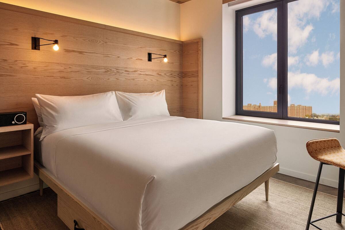 A modern bedroom with a wooden headboard, a large bed with white bedding, a nightstand with a radio, a chair, and a window with a city view.