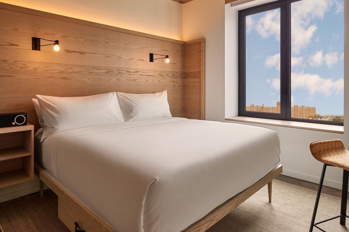 A bedroom with a wooden headboard, a bed with white bedding, a nightstand with a radio, a chair, and a window with a city view.