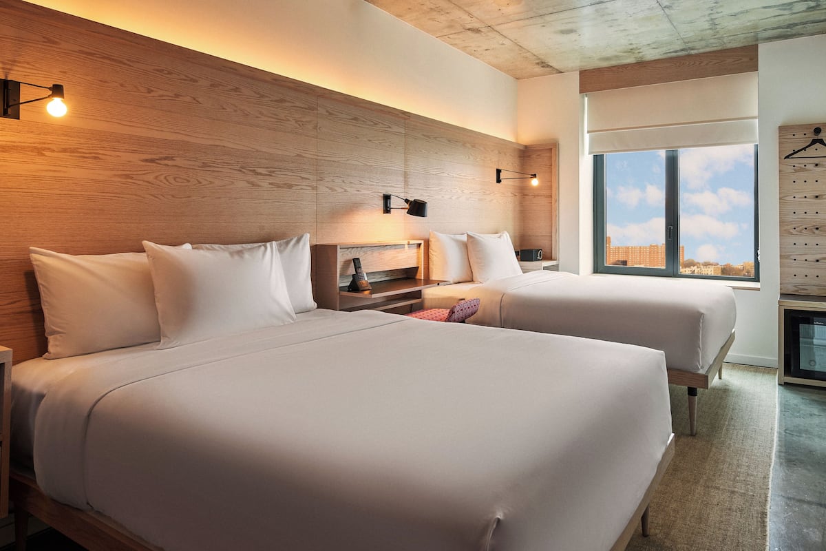 A modern hotel room with two beds, wooden headboard, bedside tables, a nightstand, a small TV, and a view of the city through large windows.