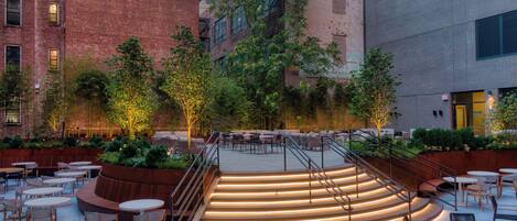 Un patio al aire libre con asientos escalonados, mesas y sillas, rodeado de vegetación y edificios.