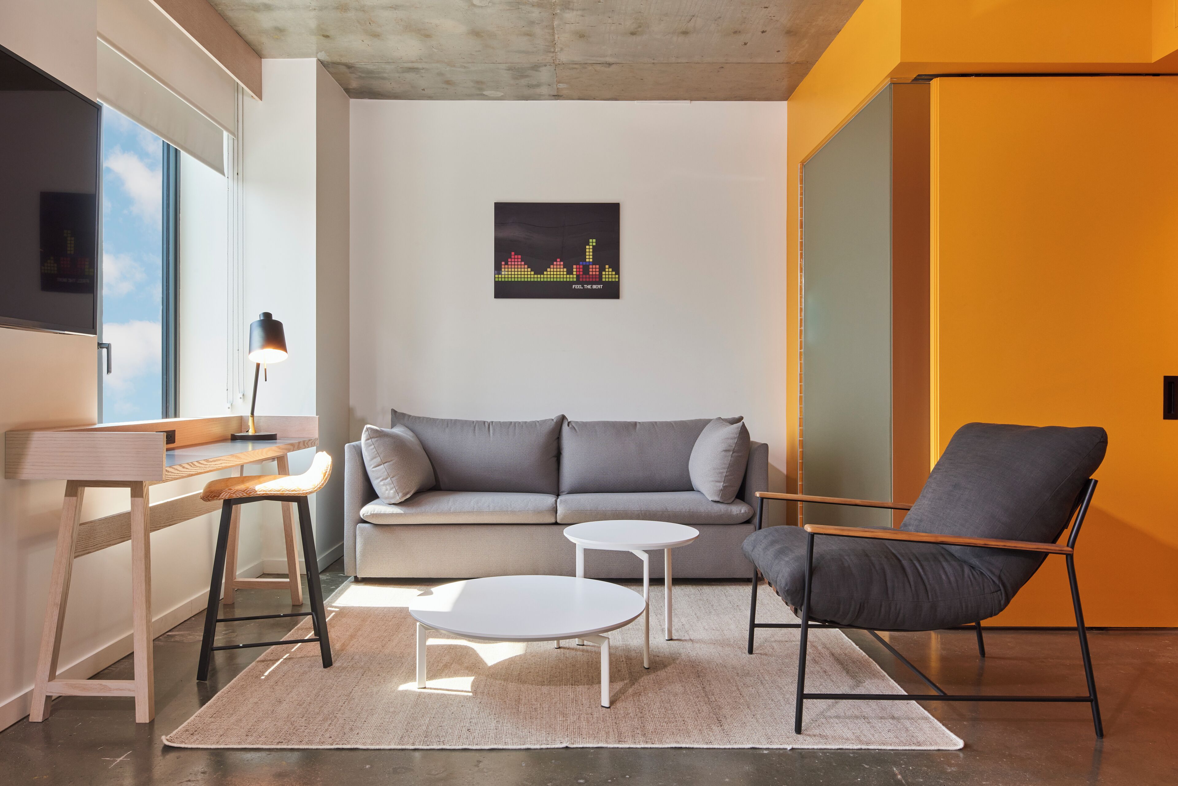 Una sala de estar moderna con un sofá gris, una mesa de centro blanca, un sillón gris y una pequeña mesa blanca. Hay un cuadro en la pared y una ventana con vistas al cielo.