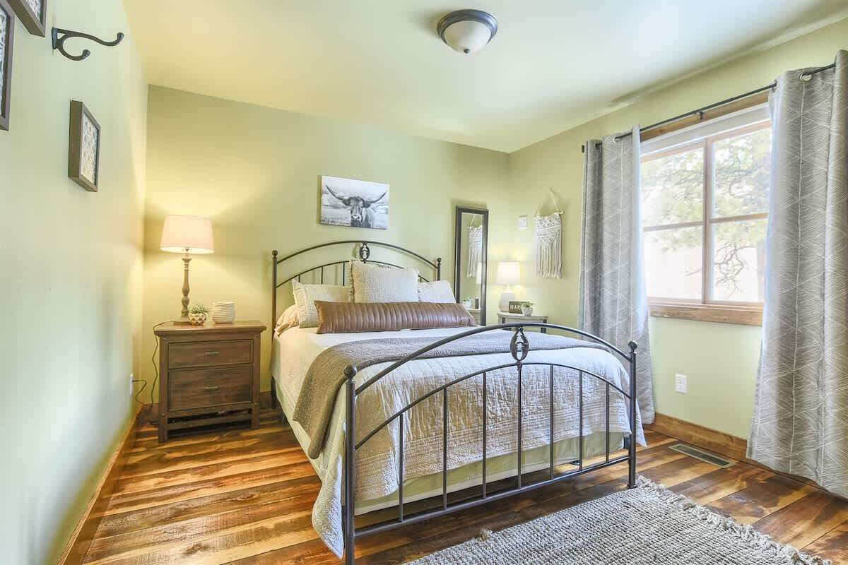 Une chambre à coucher avec un lit en métal, un plancher en bois, une table de chevet avec une lampe, une commode et une fenêtre avec des rideaux.