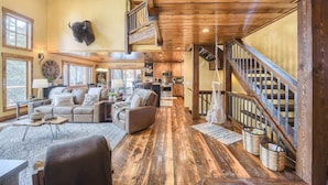 Ein geräumiges Wohnzimmer mit Holzboden, einer Sofagarnitur und einem Essbereich. Eine Treppe mit Holzwand verläuft in eine obere Ebene.