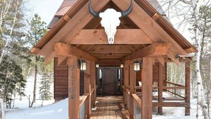 Una cabaña de madera con un imponente cráneo de toro en el techo, un corredor cubierto y un bosque nevado al fondo.