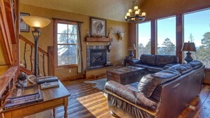 A cozy living room with a fireplace, leather sofa, and large windows offering a view of the outdoors. - Big Pine Lodge – Rustic charm, hot tub, pet friendly, and room for 12 near Terry Peak! (Lead)