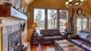 A cozy living room with leather sofas, a fireplace, and large windows offering a view of the outdoors. - Big Pine Lodge – Rustic charm, hot tub, pet friendly, and room for 12 near Terry Peak! (Lead)