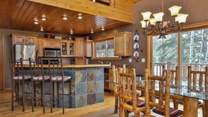 A rustic kitchen with wooden cabinets, a central island with stools, and a dining area with wooden chairs. - Deep Snow Trail Lodge (Lead)