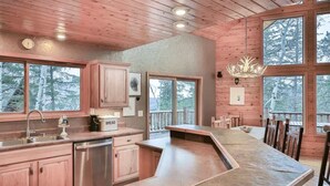 A modern kitchen with a large island, wooden cabinets, and a view of the forest through large windows. - Deep Snow Trail Lodge (Lead)