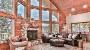 A spacious living room with a stone fireplace, large windows, and a high ceiling. - Deep Snow Trail Lodge (Lead)