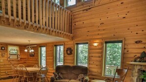 A spacious cabin with a high ceiling, wooden walls, a dining area with chairs, a sofa, and a fireplace. - Rustling Wind Haven (Lead)