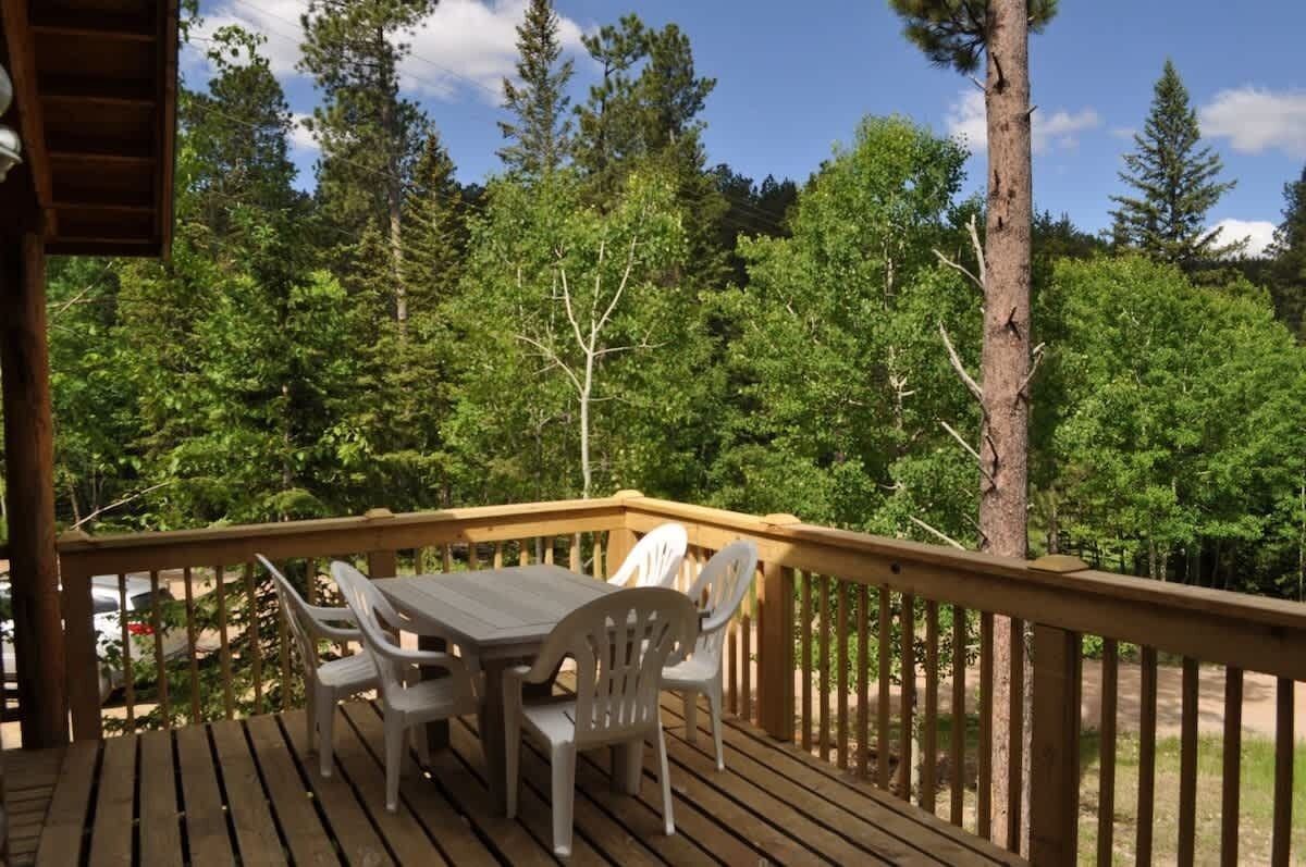 Une terrasse en bois avec une table et des chaises, entourée d’arbres verdoyants et touffus.