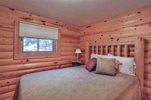 Une chambre à coucher dans un chalet en bois, avec un lit, des oreillers, une table de chevet et une fenêtre avec des stores.