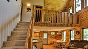 A wooden cabin interior with a staircase leading to a loft, a dining area, and a living room with a sofa and a table. - Rustling Wind Haven (Lead)