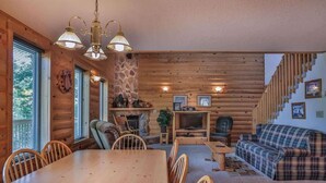 A cozy log cabin interior with a dining table, chairs, a sofa, a fireplace, and a staircase. - Rustling Wind Haven (Lead)