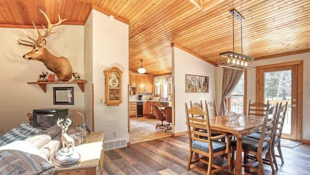 L’intérieur chaleureux d’un chalet comprend un coin repas, un salon orné d’une tête de cerf fixée au mur, et une cuisine visible à travers une porte ouverte.