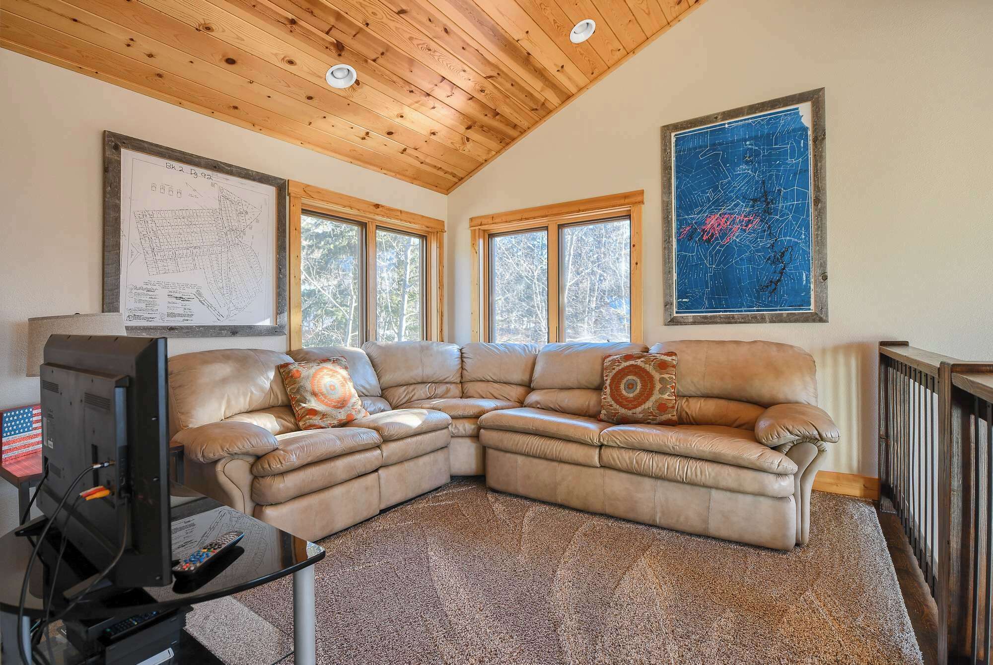 A living room with a sectional sofa, a television, and two large windows.