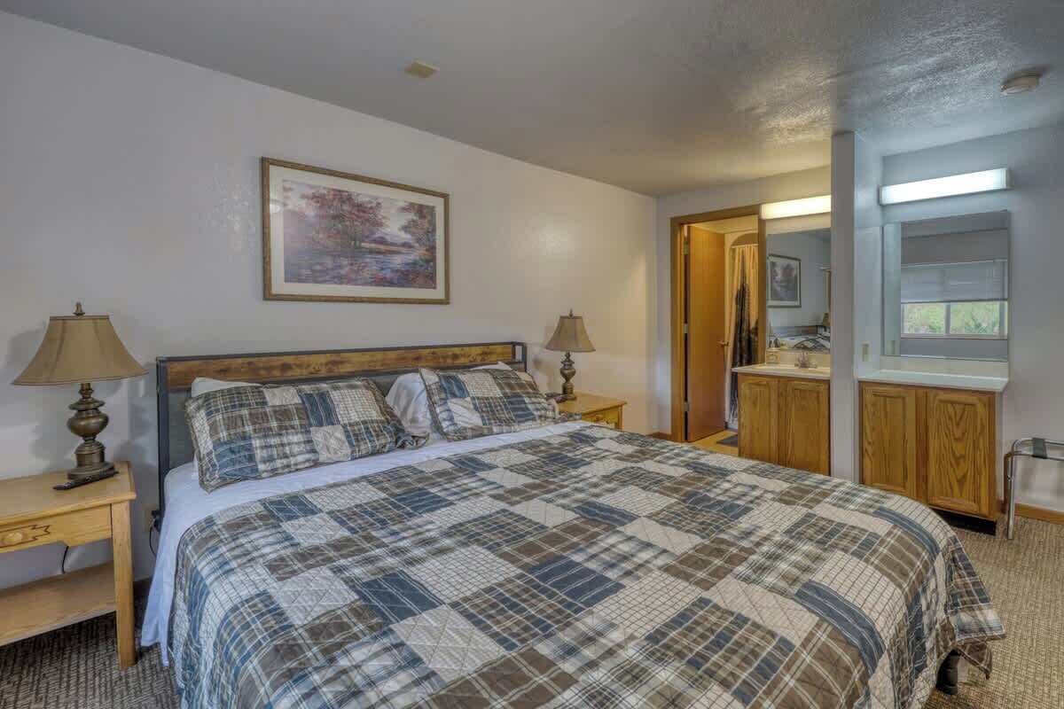 A bedroom with a large bed, two bedside tables with lamps, a wooden headboard, a framed picture on the wall, and a bathroom visible through an open door.