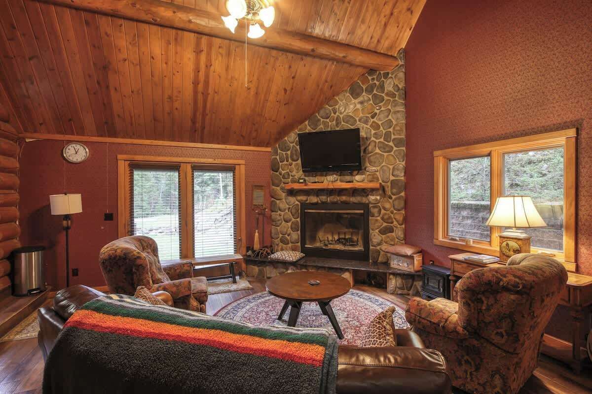 A cozy log cabin interior with a stone fireplace, leather furniture, and large windows.