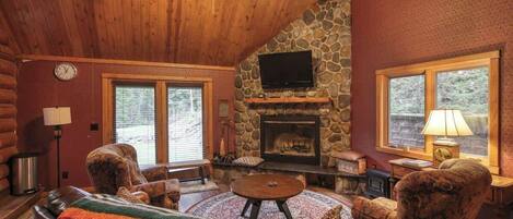 A cozy log cabin interior with a stone fireplace, leather furniture, and large windows.