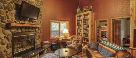 A cozy living room with a stone fireplace, a television mounted on the wall, a comfortable sofa, a bookshelf, and a large window providing natural light.