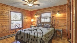 A wooden cabin bedroom with a bed, two windows, a ceiling fan, and a nightstand. - Whispering Grace Retreat (Rapid City)