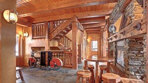 Um interior rústico com lareira de pedra, escada de madeira e uma mesa redonda com bancos.