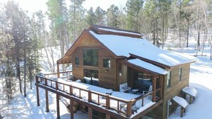 A wooden cabin with a deck, surrounded by trees and covered in snow.