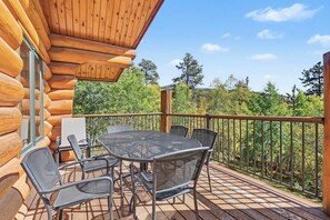 Une terrasse en bois, agrémentée d’une table et de chaises, entourée d’une rambarde en métal, et offrant une vue sur une zone boisée.