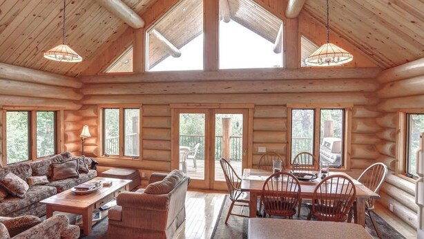 A log cabin interior with a dining area, sofa, and large windows. - Dakota Log Cabin – Rustic charm, amazing views, wildlife watching & cozy family spaces! (Lead)