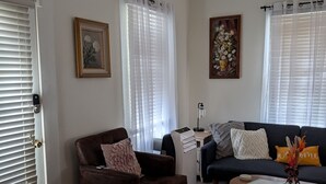 A living room with a brown leather armchair, a dark sofa, a coffee table, and a rug. There are two windows with white curtains, a door with a handle, and a painting on the wall. - Cozy, Cute, Private Ozark Suite 5 Mins to Hospital (Ozark)