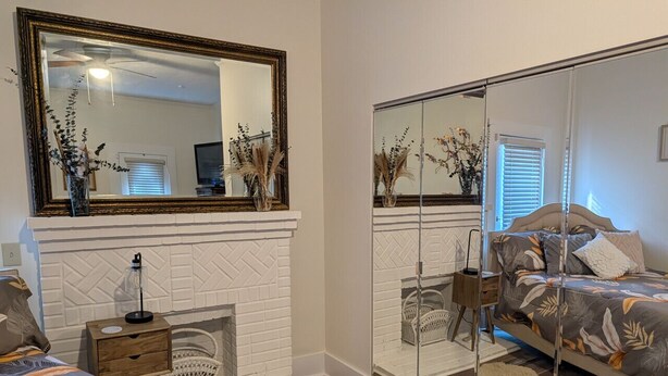 A bedroom with a fireplace, a bed with floral bedding, a nightstand, and a mirror with a gold frame. - Cozy, Cute, Private Ozark Suite 5 Mins to Hospital (Ozark)