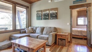A cozy room with a sofa, a wooden table, and a bed. There are large windows and a sign that says "Carrabassett Valley." - Après Powder Haus – Walk to the slopes!  Hot tub, pet friendly and game garage (Lead)