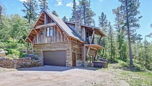A rustic cabin with a stone foundation, a garage, and a large deck overlooking a forested area. - Après Powder Haus – Walk to the slopes!  Hot tub, pet friendly and game garage (Lead)