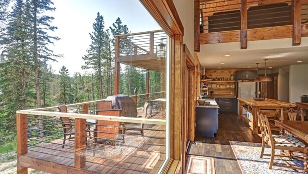 Une vaste terrasse en bois avec coin salon extérieur, un espace cuisine doté de meubles en bois et une grande fenêtre donnant sur une zone boisée.