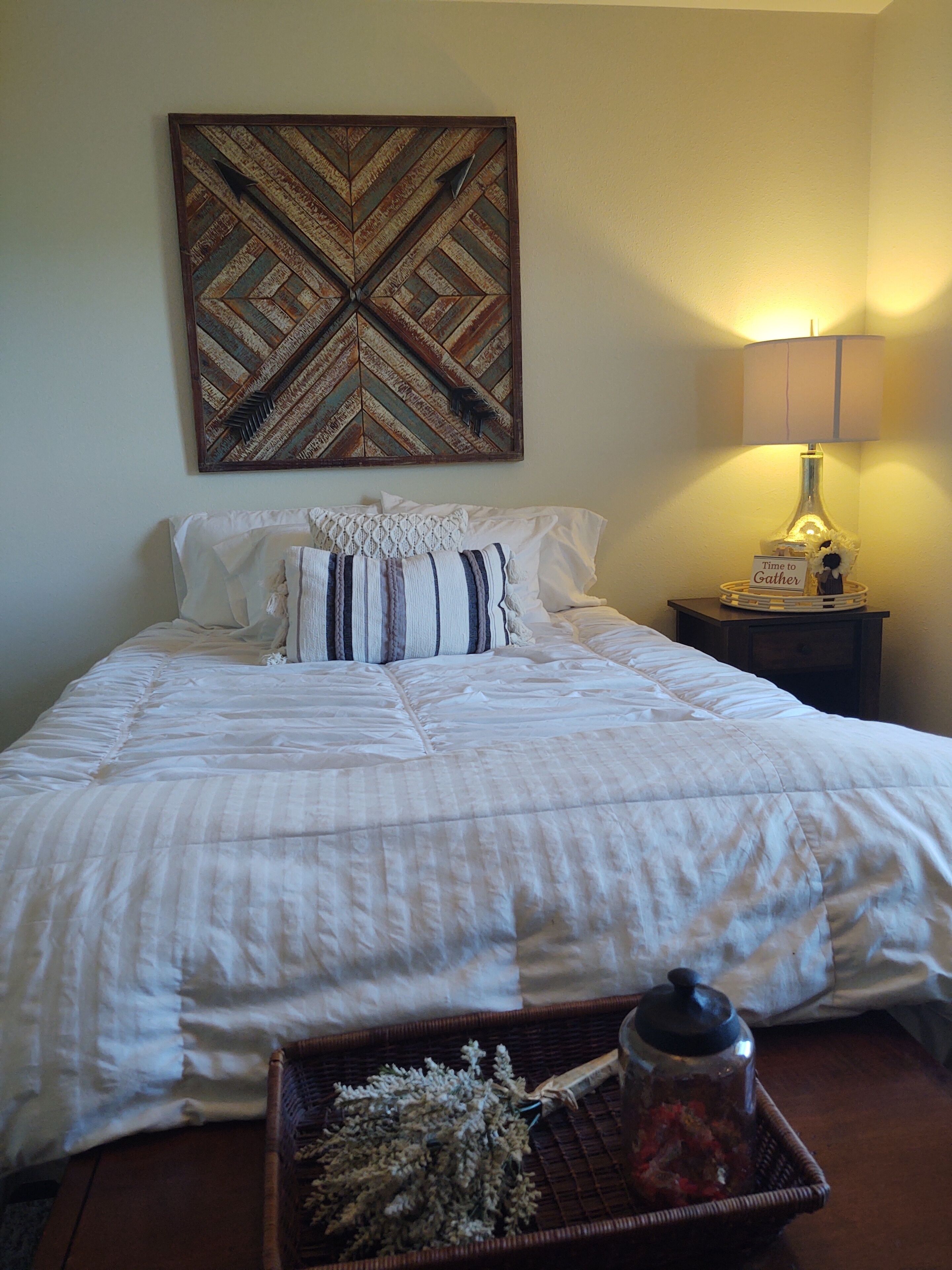 A neatly made bed with a striped comforter and pillows, a decorative wall piece, a bedside lamp, and a tray with dried flowers and a jar.